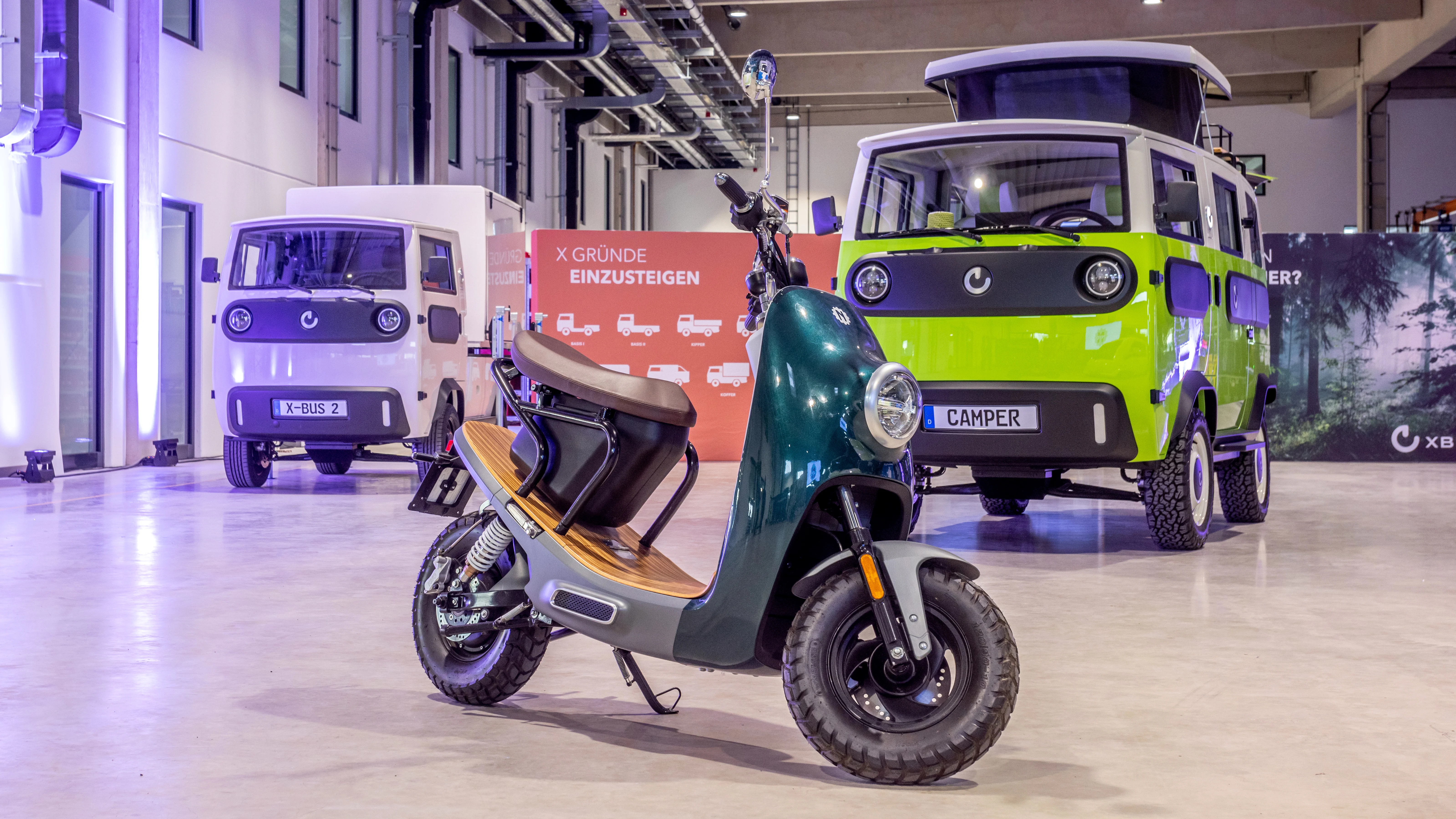 Modern electric scooter in foreground with compact green camper van and white X-Bus 2 vehicle in a showroom.