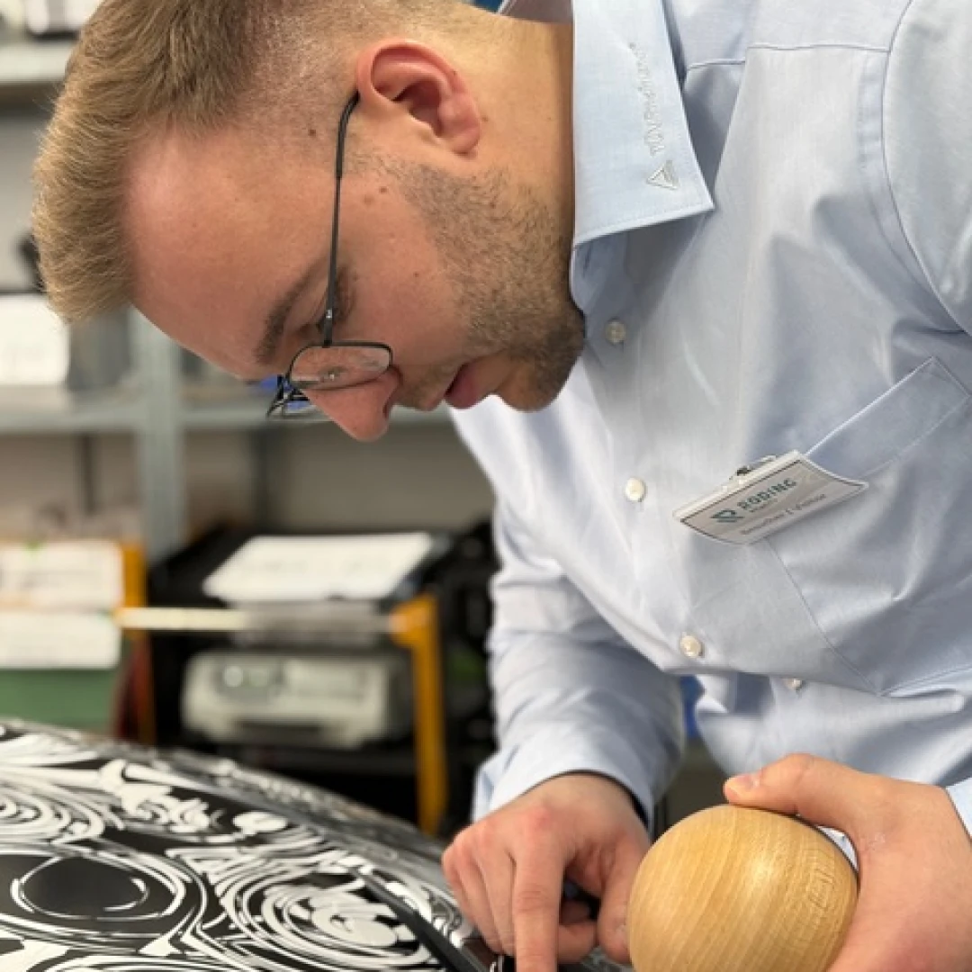 Man in glasses closely inspecting black and white patterned surface while holding a wooden tool.