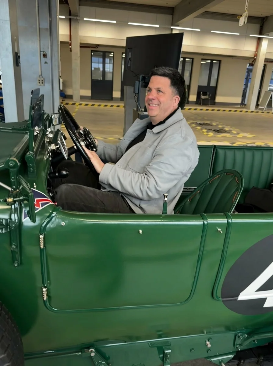 Mann sitzt lächelnd in einem grünen Oldtimer-Auto in einer Halle.