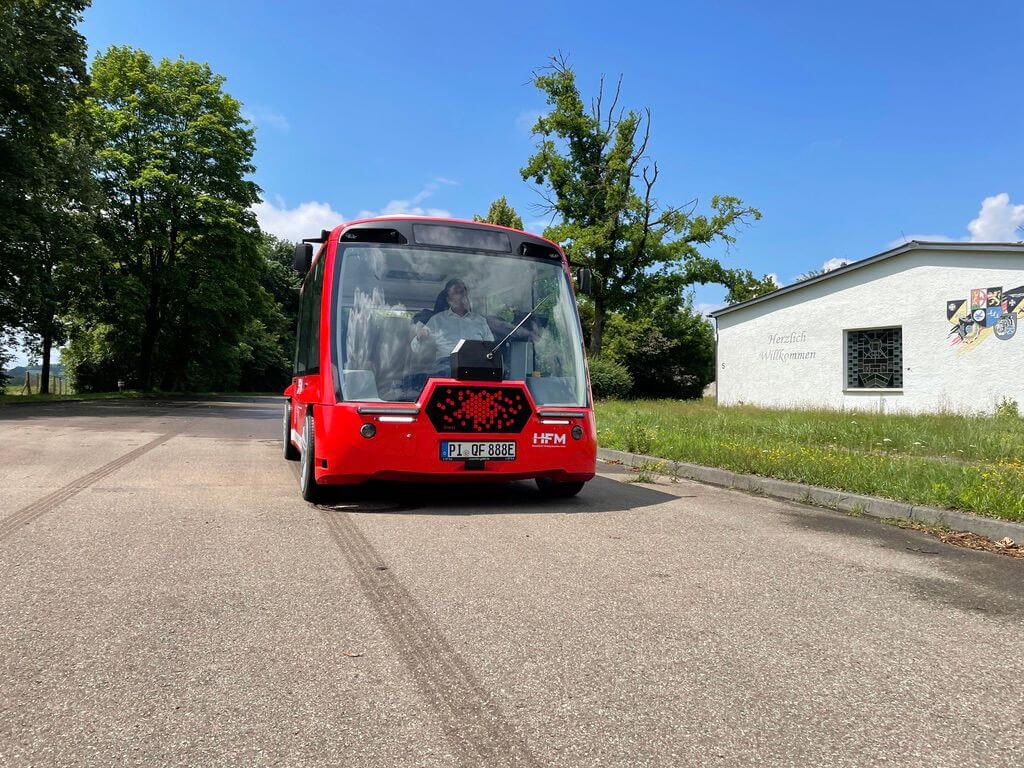 Rotes autonom fahrendes Fahrzeug auf einer Straße mit Bäumen und einem Gebäude in der Nähe.