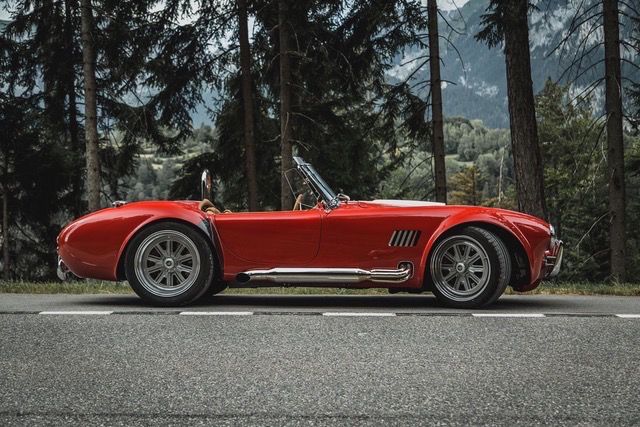 Red vintage convertible sports car parked on a forest road with trees in the background.