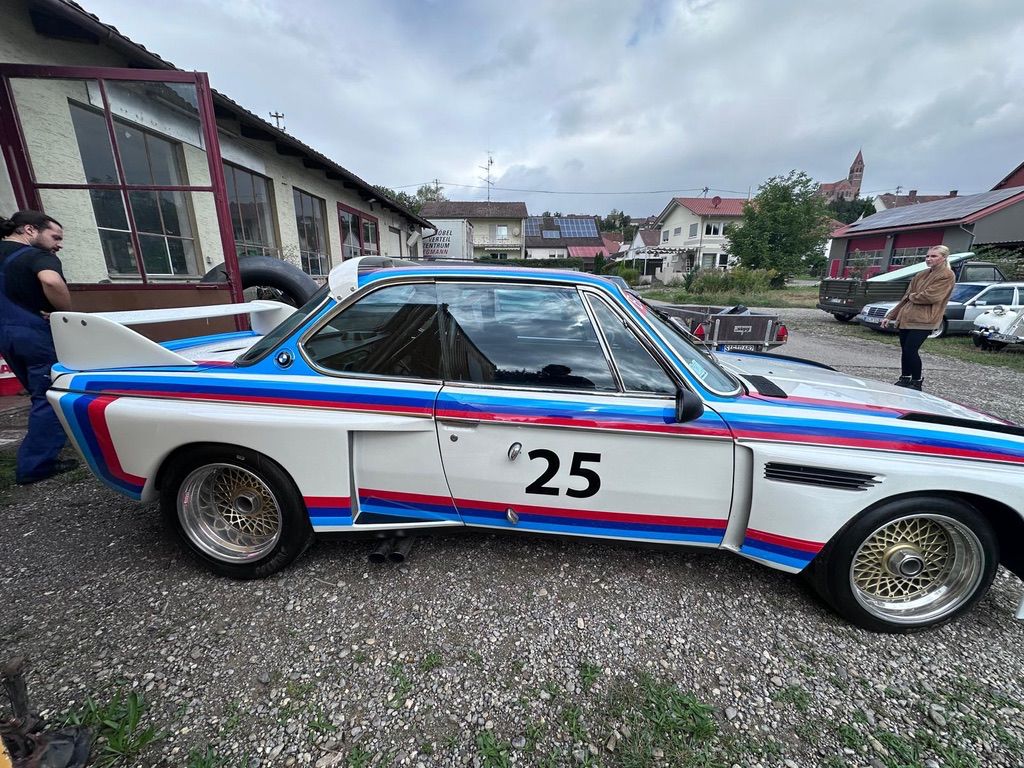 Side view of a white vintage race car with the number 25 and red, blue, and light blue striping parked on a gravel surface.