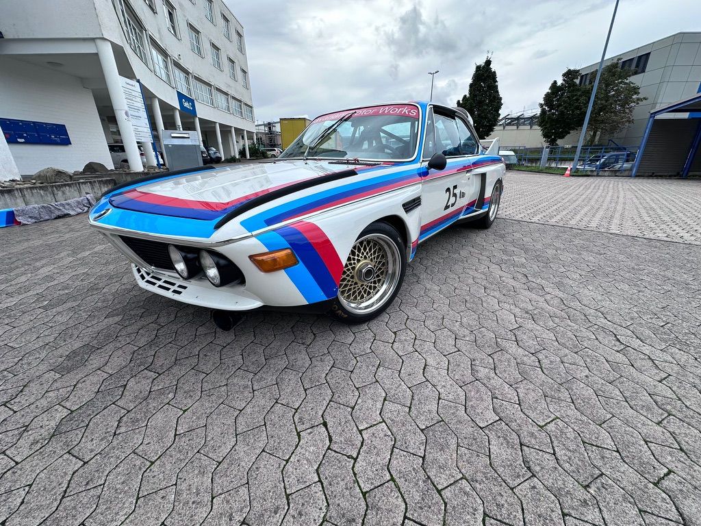 White vintage race car with blue and red racing stripes parked on pavement outside commercial buildings.