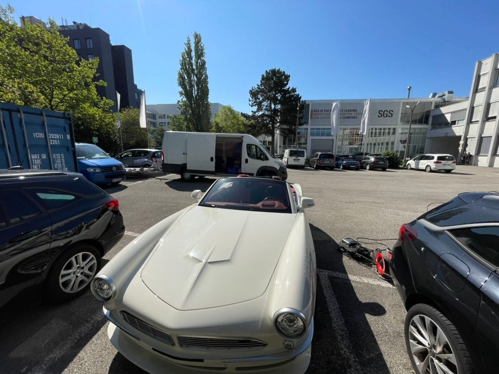White vintage convertible car parked in a lot between two black cars with a white van and SGS building in the background.