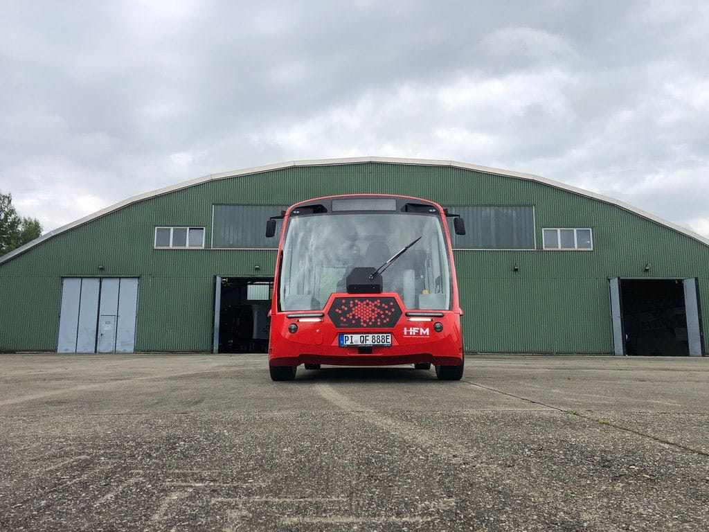 Front view of a small red autonomous vehicle parked on concrete in front of a large green industrial building under a cloudy sky.