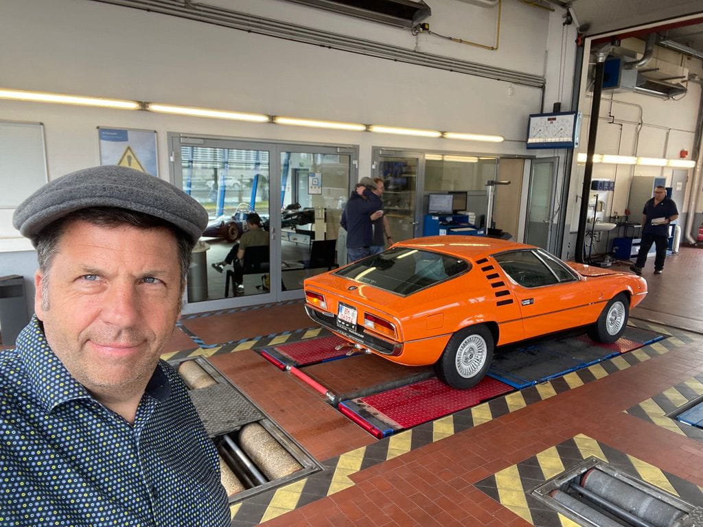 Man in a flat cap taking a selfie inside a car service garage with a bright orange vintage sports car on roller testing equipment in the background.