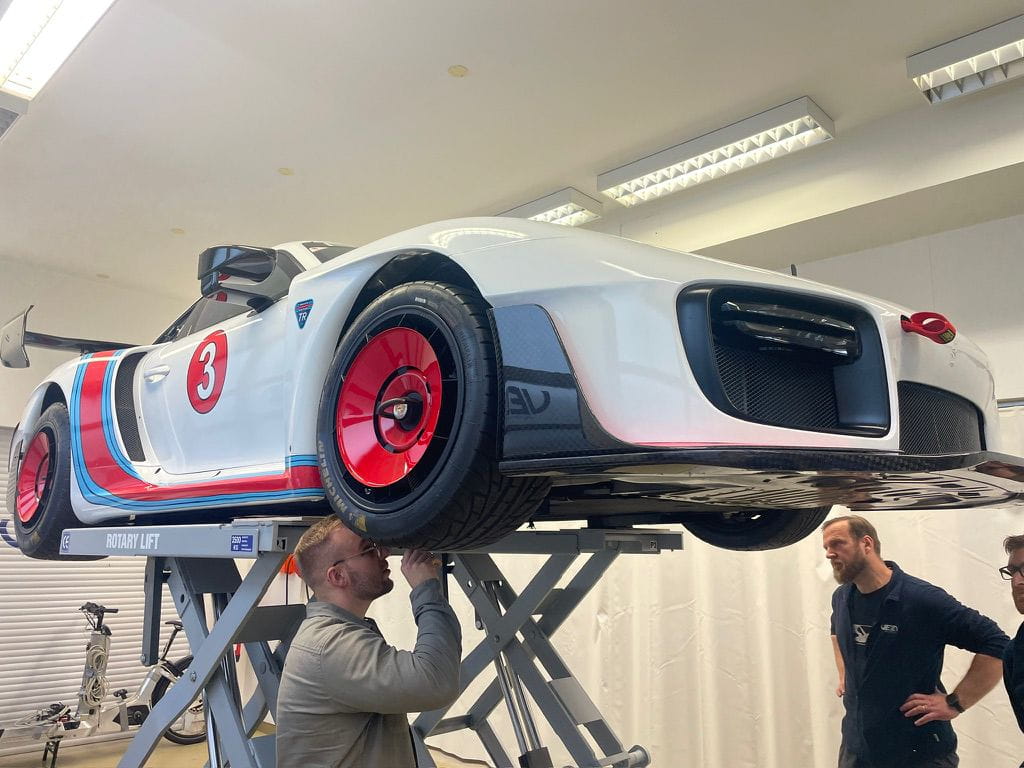 White race car with red wheels and number 3 on the side, elevated on a rotary lift with three men inspecting it in a workshop.