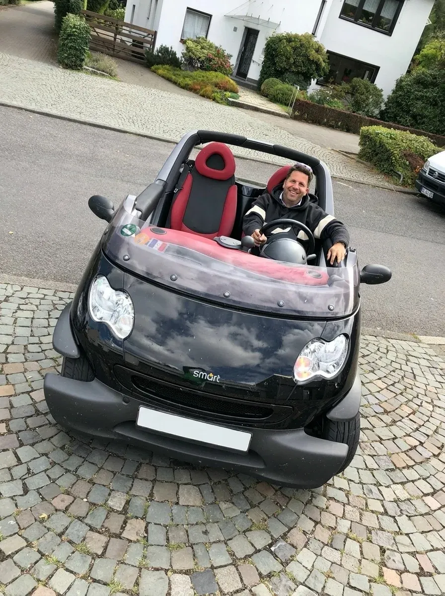 Smiling man sitting in a black Smart convertible car with red and black seats, parked on a cobblestone street in front of a white house.