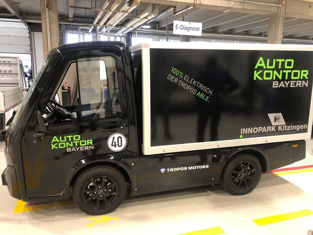Compact black electric delivery vehicle with green text 'Auto Kontor Bayern' and '100% elektrisch. Der Tropos able.' inside an industrial indoor garage.