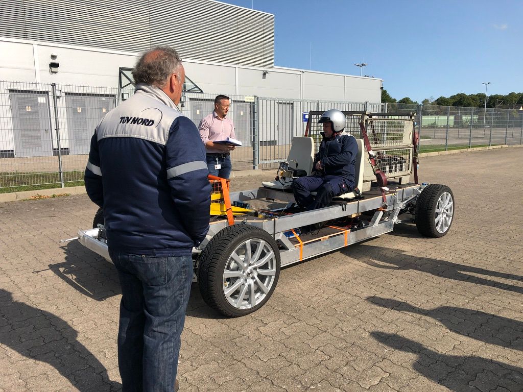 Three men near an unfinished car chassis with two seats, one man seated wearing a helmet, outdoors near a fenced industrial area.