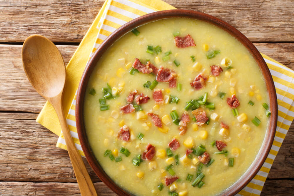 a bowl of sweet corn chowder resting next to a wooden soup spoon
