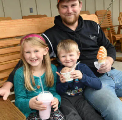 Man sitting on a wooden bench smiling with a young girl holding a pink milkshake and a young boy holding a cup of ice cream.