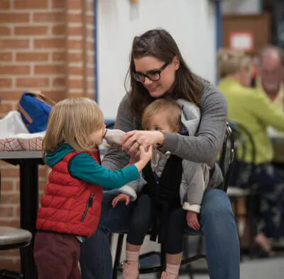 Woman wearing glasses sitting and helping a young child drink from a bottle while another child looks on.