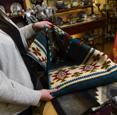 Person holding and displaying a colorful woven rug with geometric patterns in a shop filled with pottery and other items.