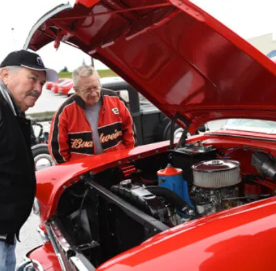 Two older men examining the engine of a red vintage car with its hood open.