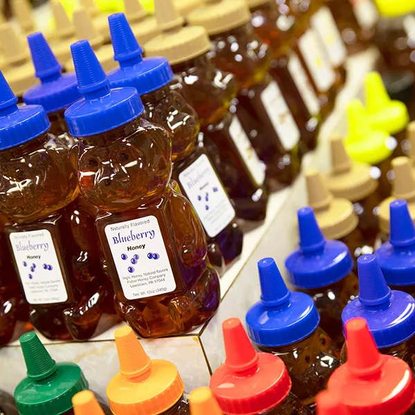 Rows of bear-shaped honey bottles with blue, beige, yellow, green, orange, and red caps, labeled blueberry honey.