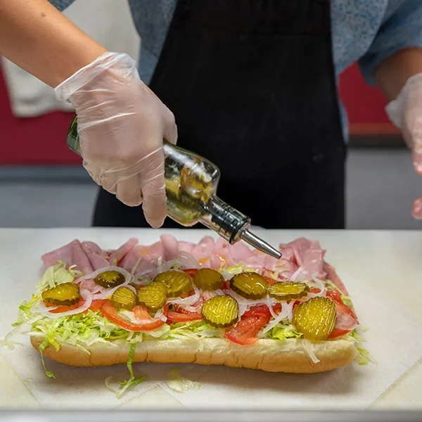 Person wearing gloves drizzling oil over a sandwich with ham, lettuce, tomato, onions, and pickles on a white sub roll.