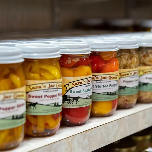 Shelf with jars of Sara's Jar Goods containing colorful stuffed peppers and other preserved foods.
