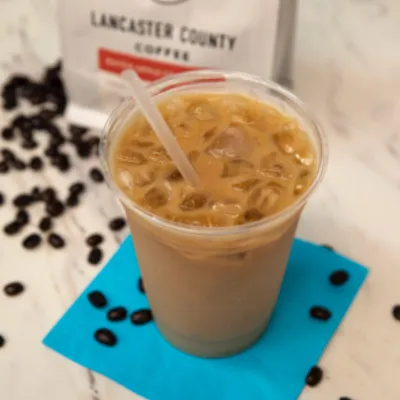 Iced coffee in a plastic cup with a straw on a blue napkin, coffee beans scattered around.