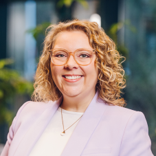 Smiling woman with curly blonde hair and glasses wearing a light purple blazer and necklace.