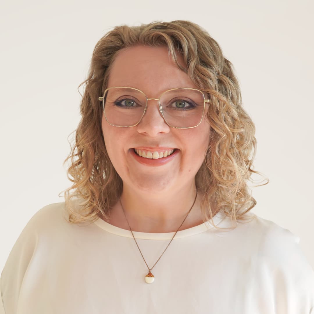 Smiling woman with curly blonde hair, wearing glasses, a white top, and a necklace with a round pendant.