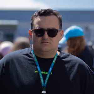Fraser Singleton wearing black sunglasses and a black shirt with a blue lanyard around his neck, blurred crowd and building in the background.