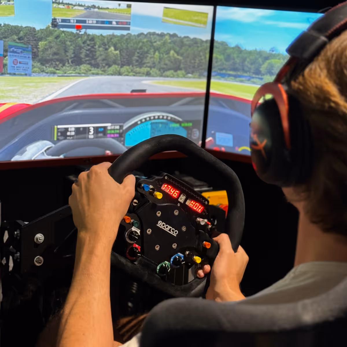 Person wearing headphones gripping a Sparco racing simulator steering wheel with a racing game displayed on a triple screen setup.
