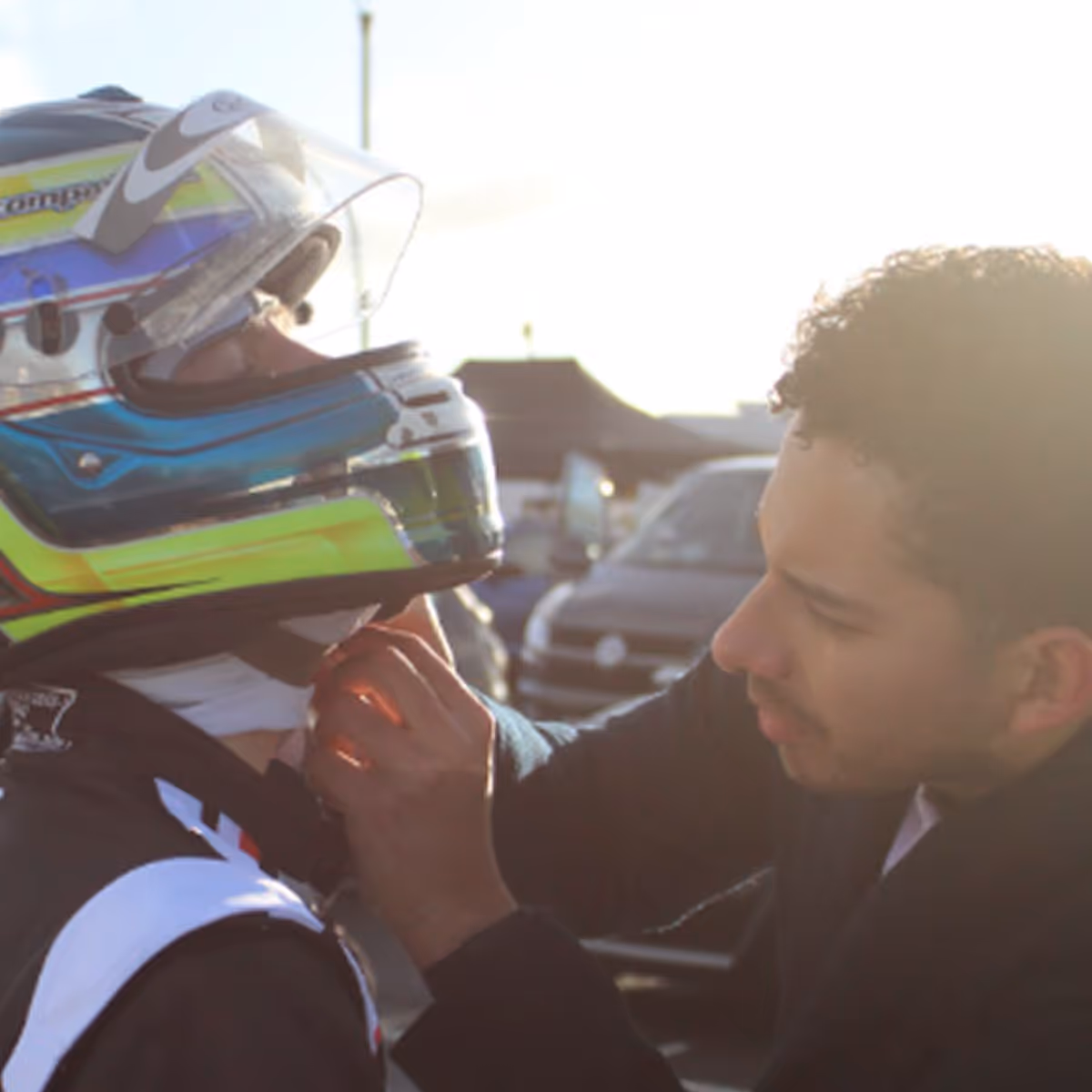 Man helping a person wearing a colorful racing helmet adjust their suit outdoors with vehicles in the background.