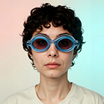 Person with short curly hair wearing blue swimming goggles and a white shirt against a gradient background.