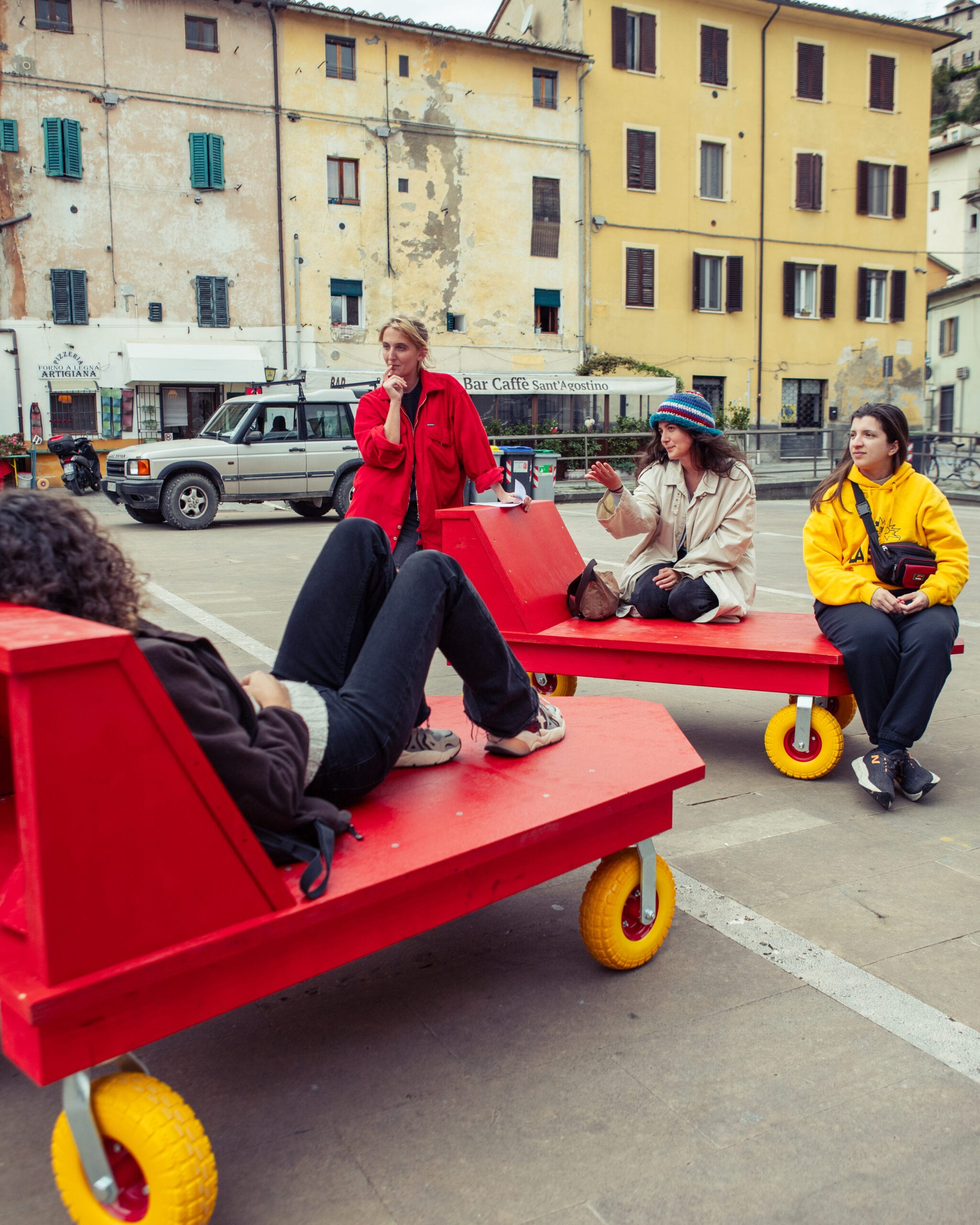 Persone sedute e sdraiate su sedute rosse su ruote in una piazza circondata da edifici