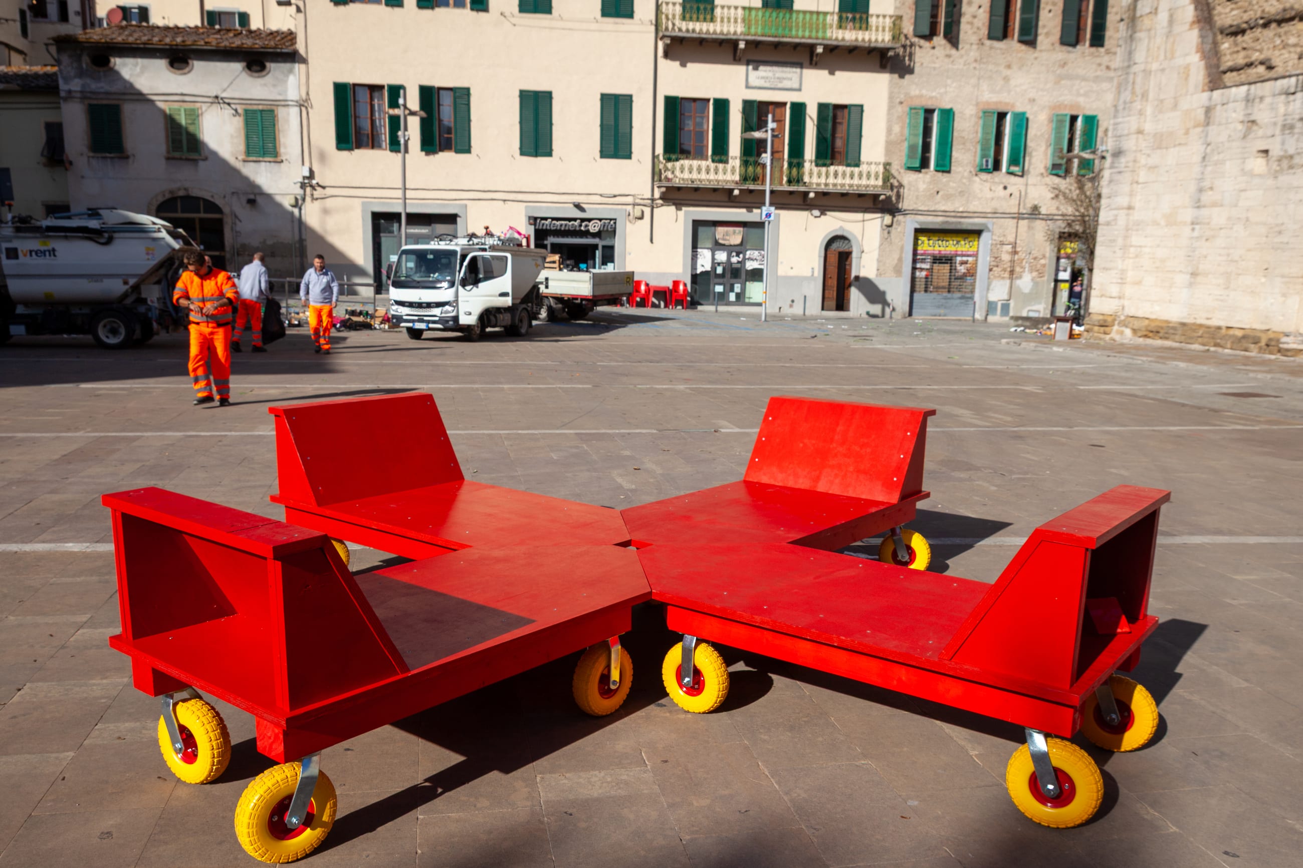 Sedute rosse su ruote gialle accostate in una piazza, davanti a una fila di edifici