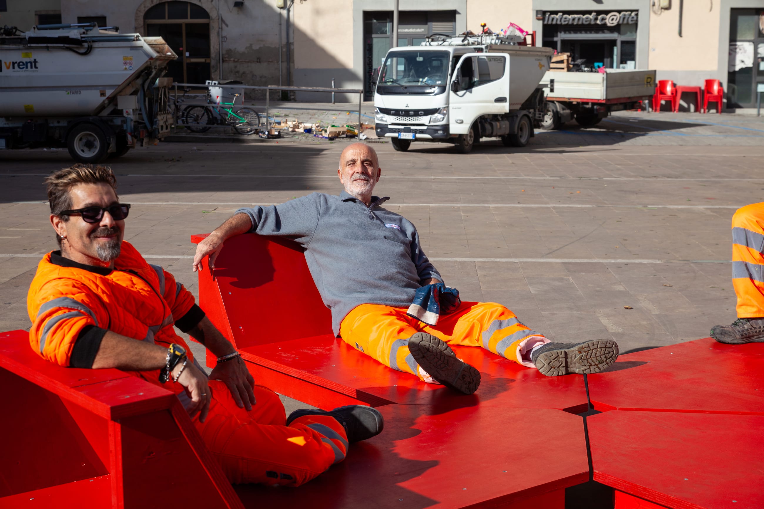 Due persone in abiti da lavoro sedute sui moduli rossi, in una piazza con dei furgoni