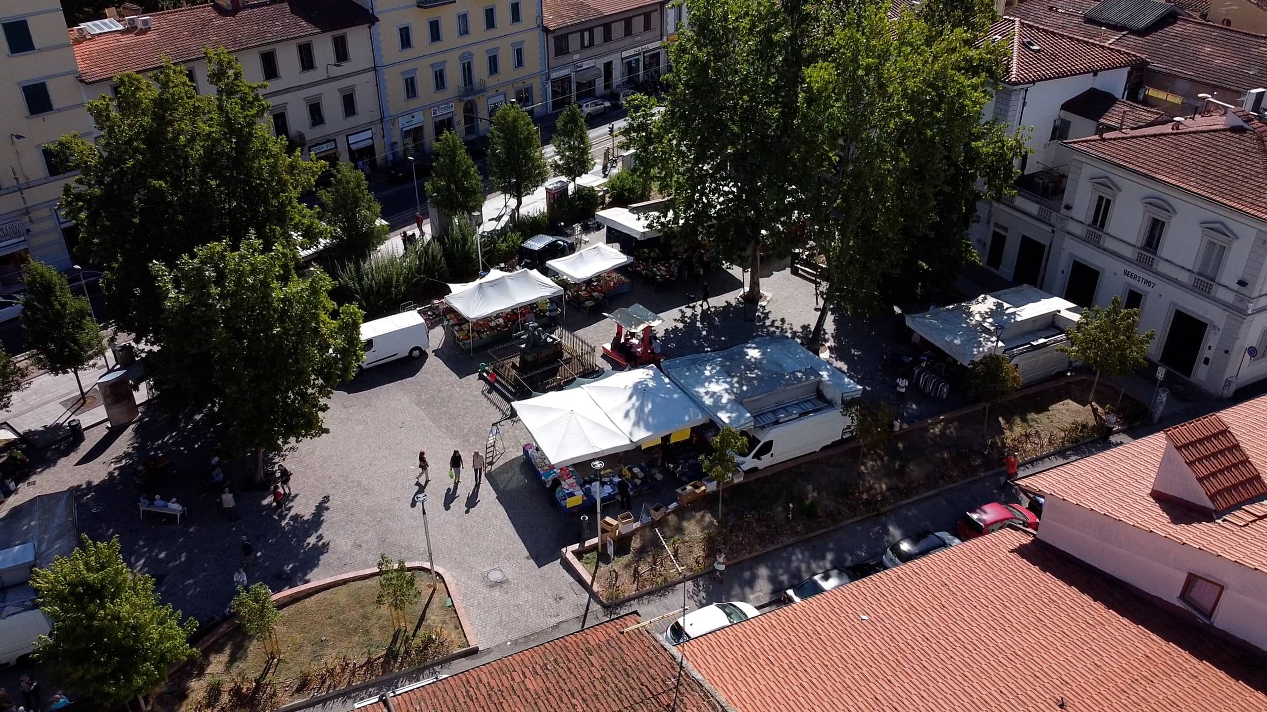 Vista aerea di una piazza alberata con i gazebo di un mercato, tra tetti e edifici