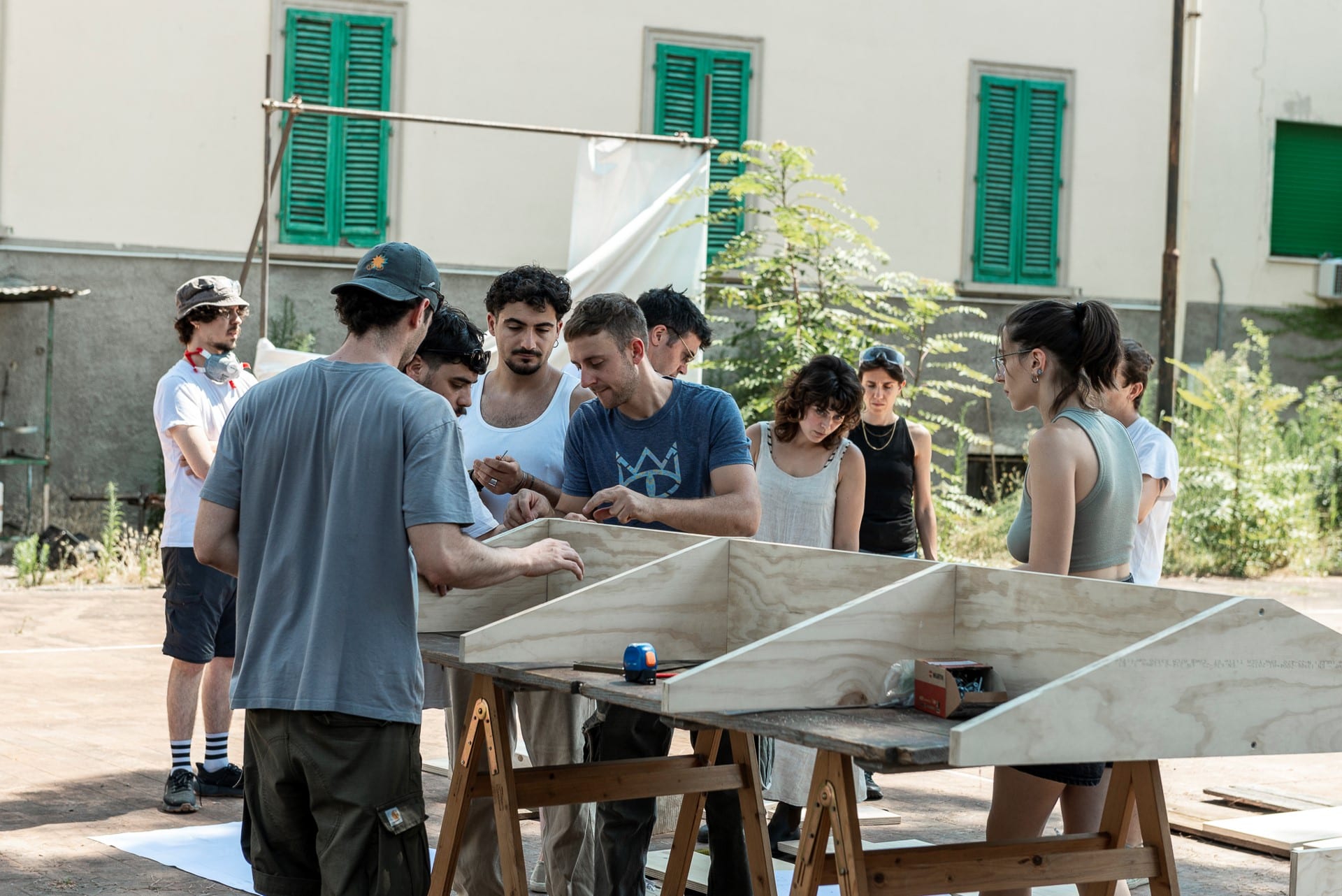 Gruppo al lavoro su pannelli in legno appoggiati su cavalletti, in un cortile