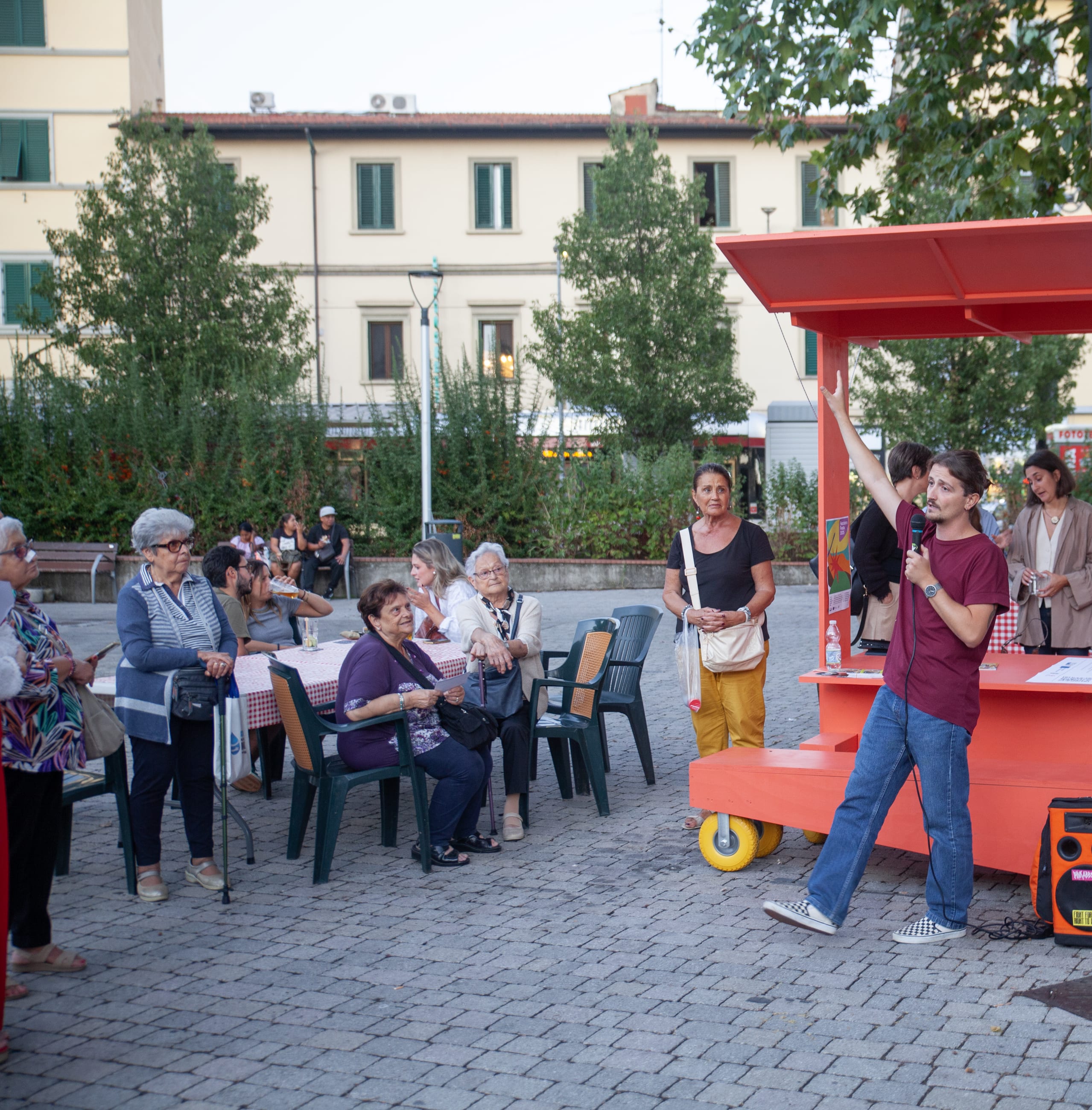 Persona parla al microfono accanto a una struttura arancione su ruote, davanti al pubblico