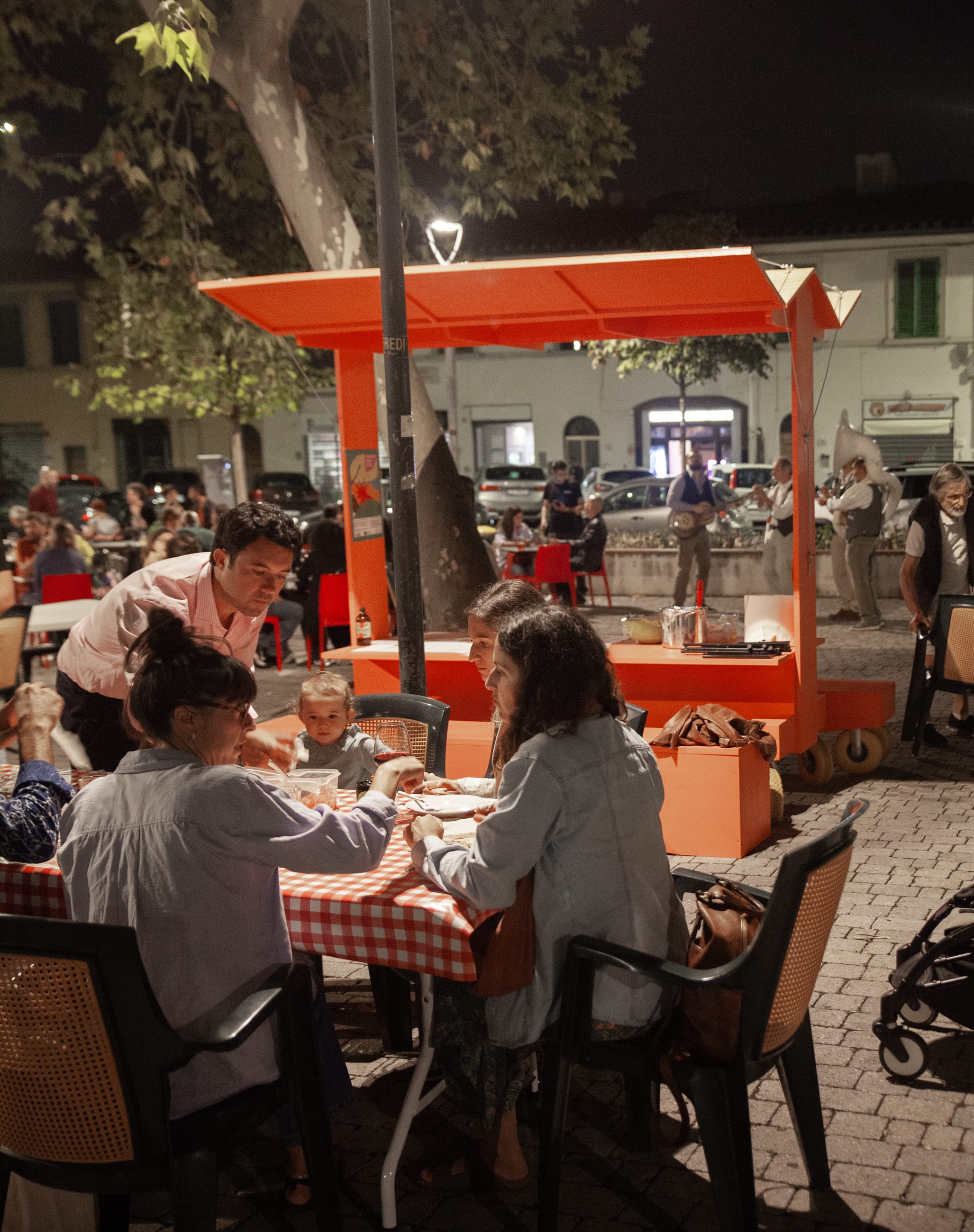 Persone cenano di sera a tavoli con tovaglie a quadri, accanto alla struttura arancione