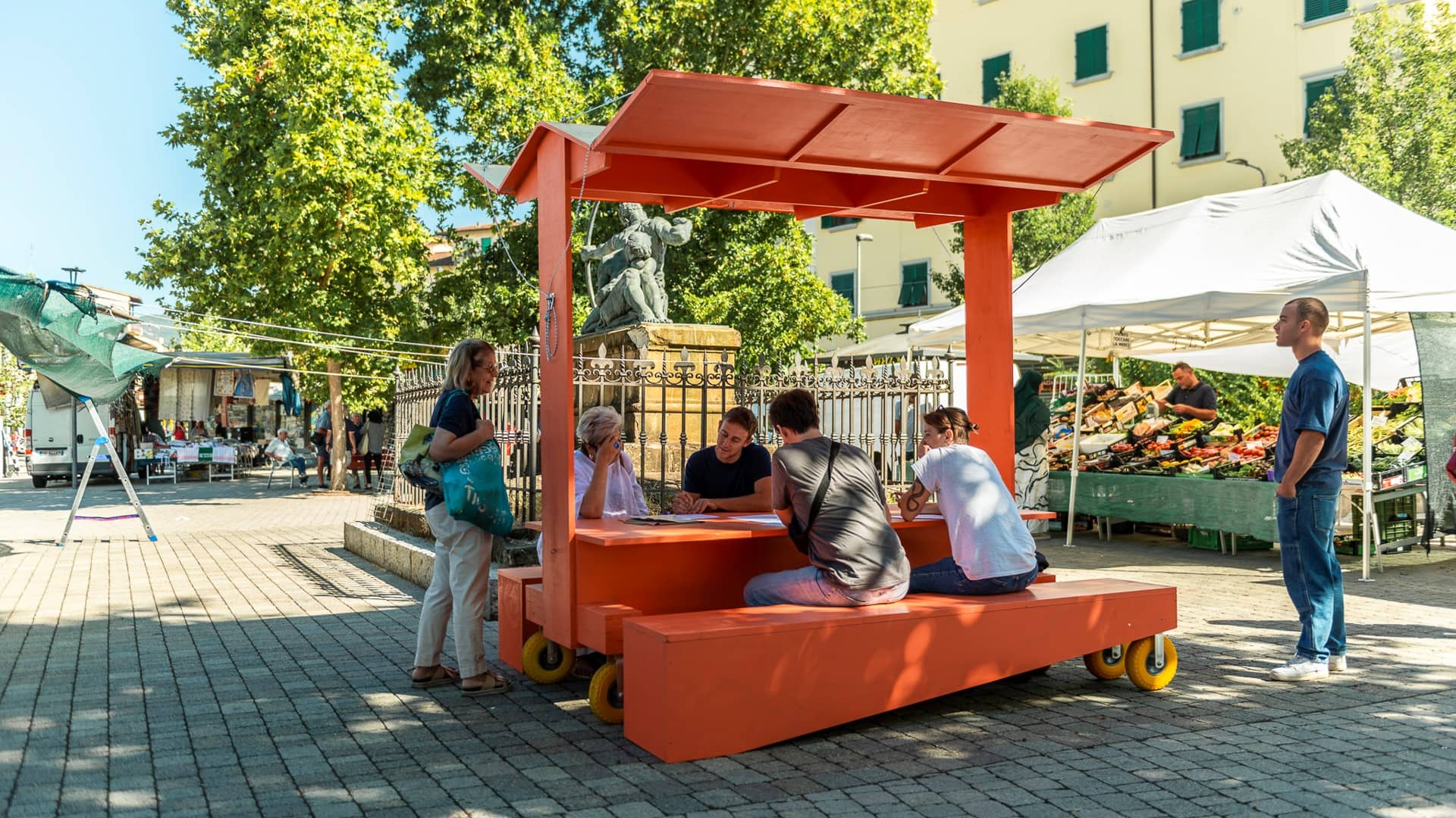 Tavolo arancione su ruote con tettoia, attorno persone sedute in una piazza con mercato