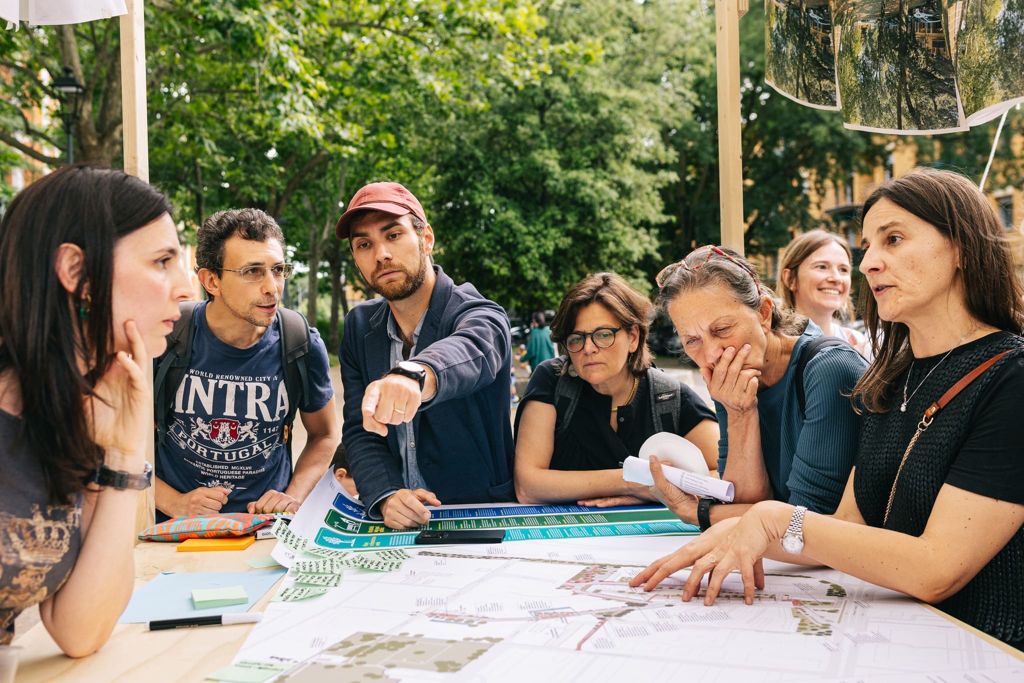 Persone discutono attorno a un tavolo coperto di mappe e disegni, sotto un padiglione