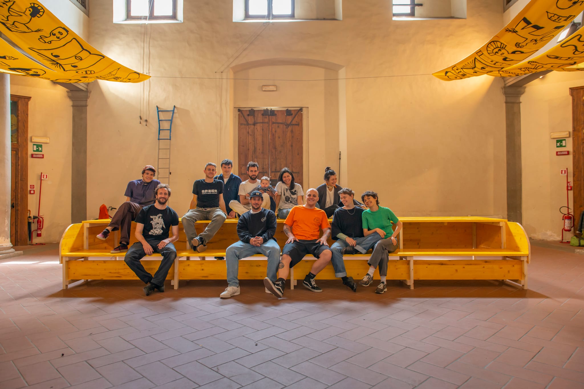 Gruppo in posa su una lunga panca in legno giallo in un salone storico