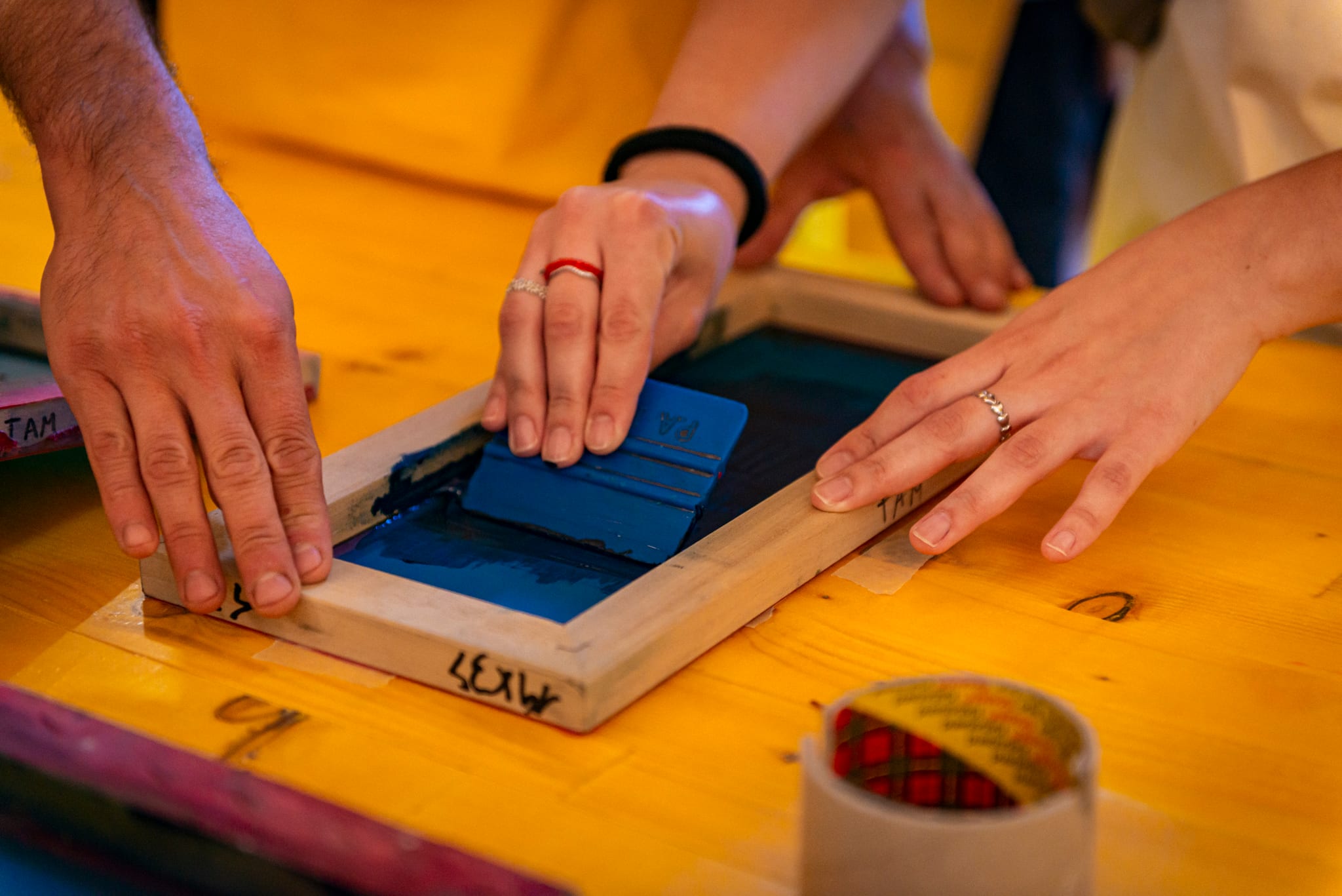 Mani stendono inchiostro blu su un telaio serigrafico appoggiato su un tavolo giallo