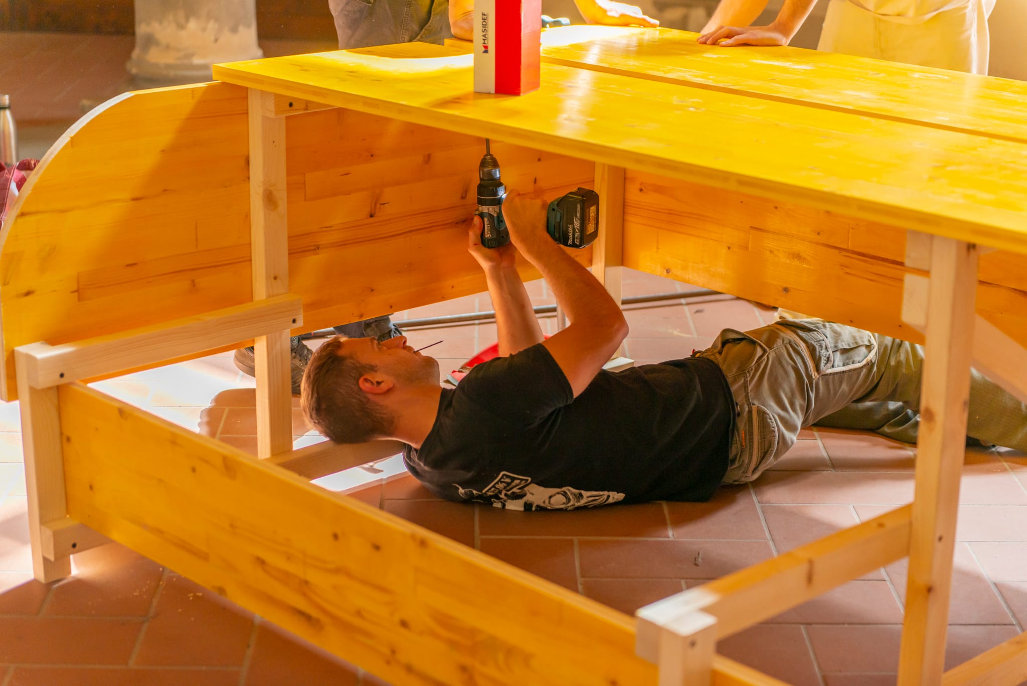 Persona sdraiata a terra che avvita con un trapano la struttura in legno di un tavolo