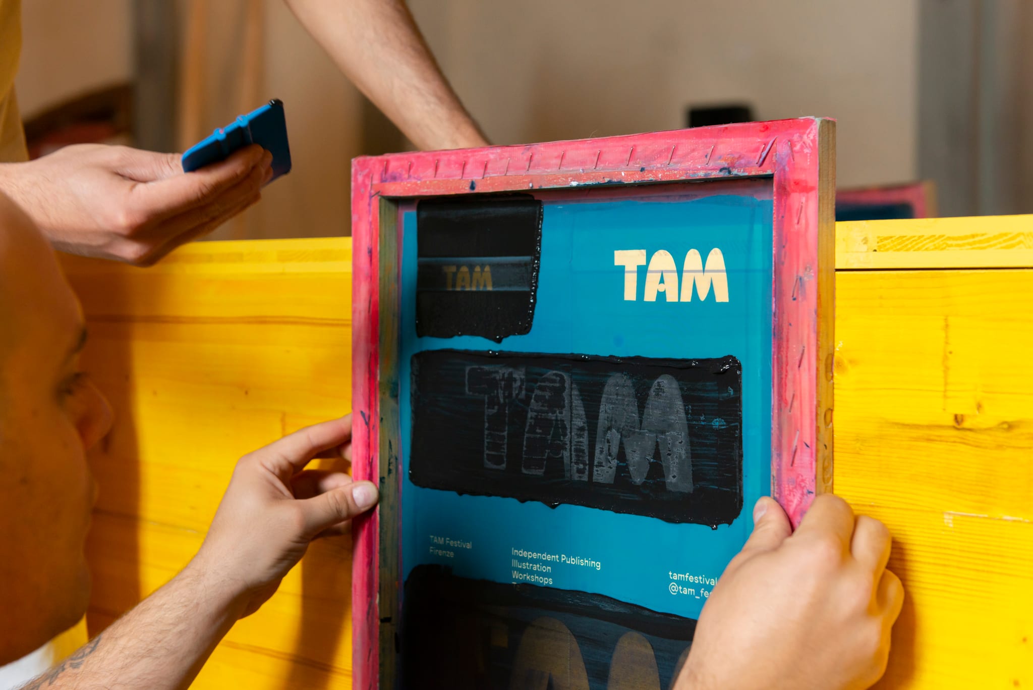 Mani sollevano un telaio serigrafico da una stampa appena realizzata su un tavolo giallo