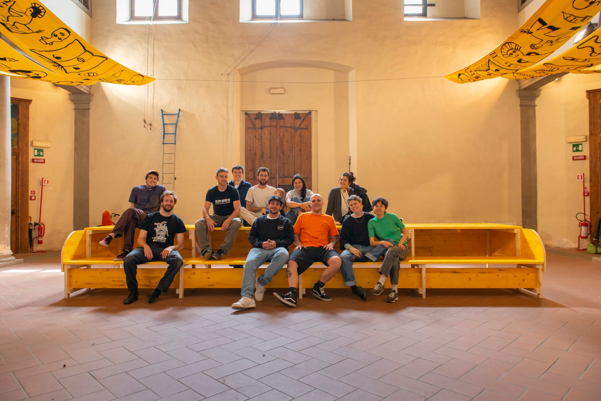 Gruppo seduto su una lunga panca in legno giallo al centro di un salone storico