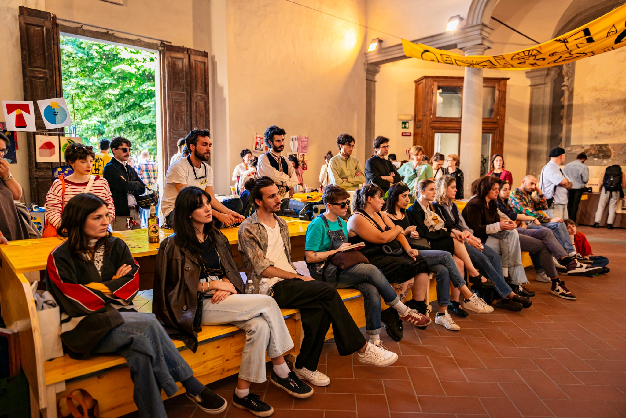 Pubblico seduto su panche in legno giallo durante un incontro in un salone storico