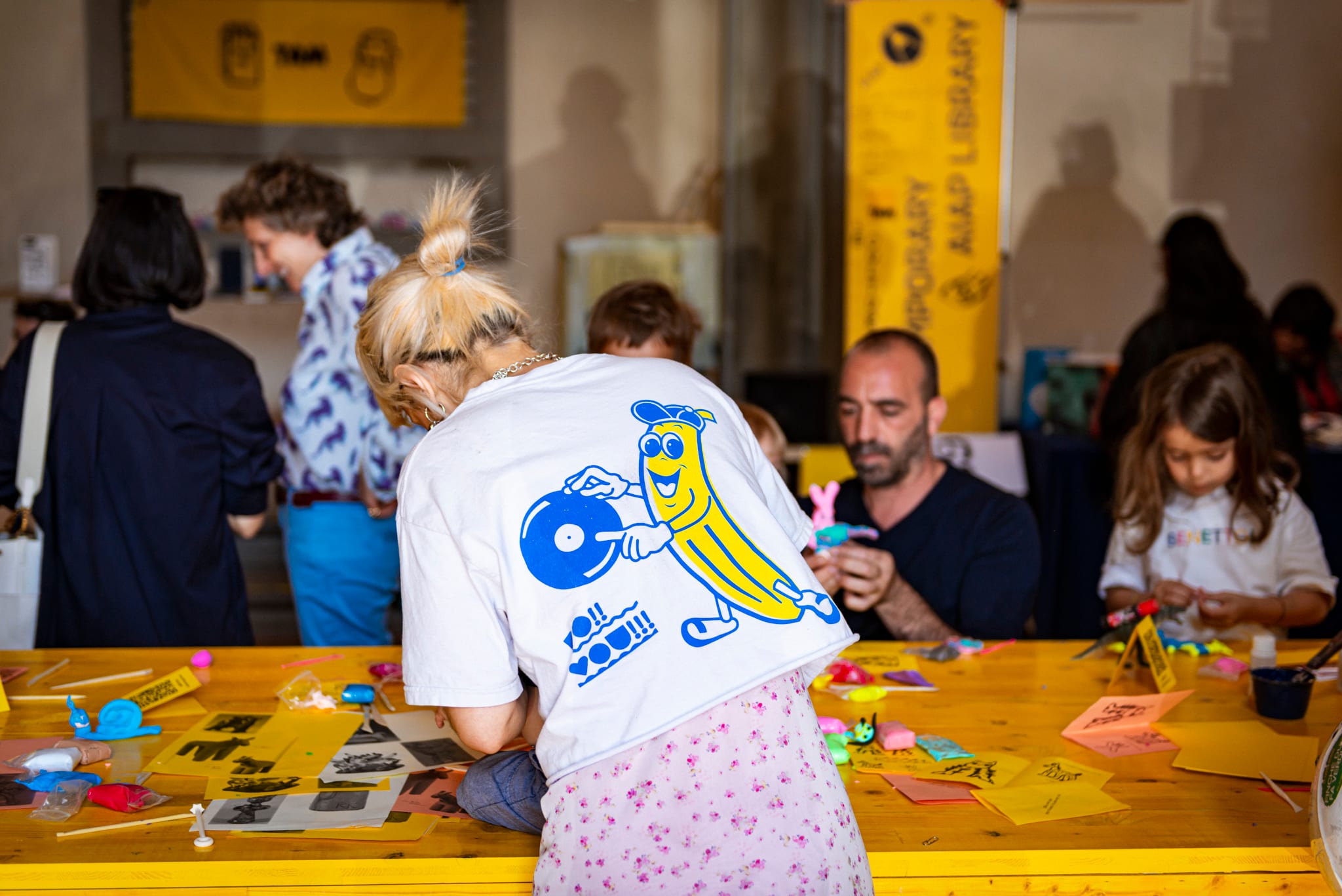 Adulti e bambini a un tavolo giallo pieno di carte e materiali per attività creative