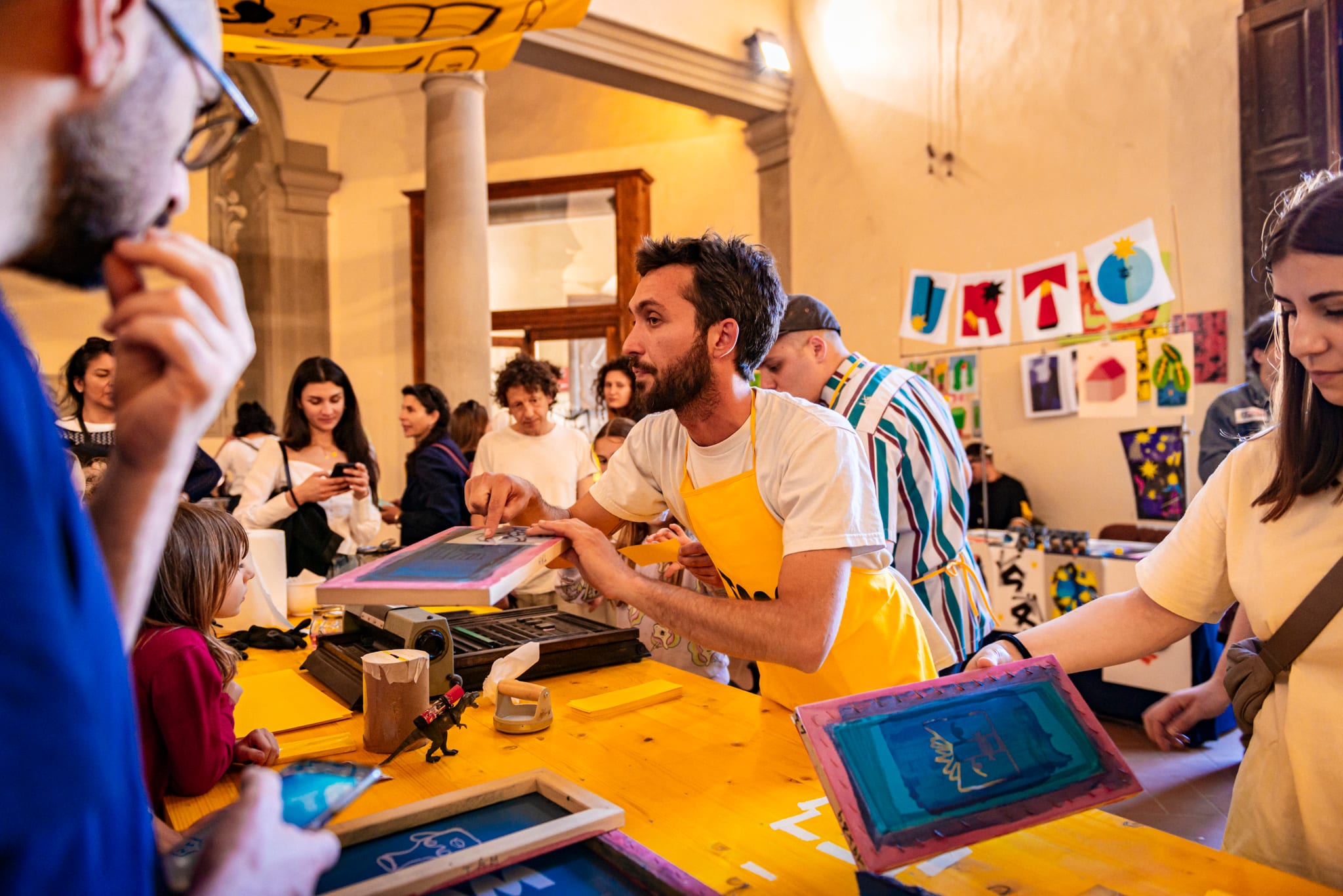 Persona con grembiule giallo guida una stampa serigrafica, tra persone e bambini
