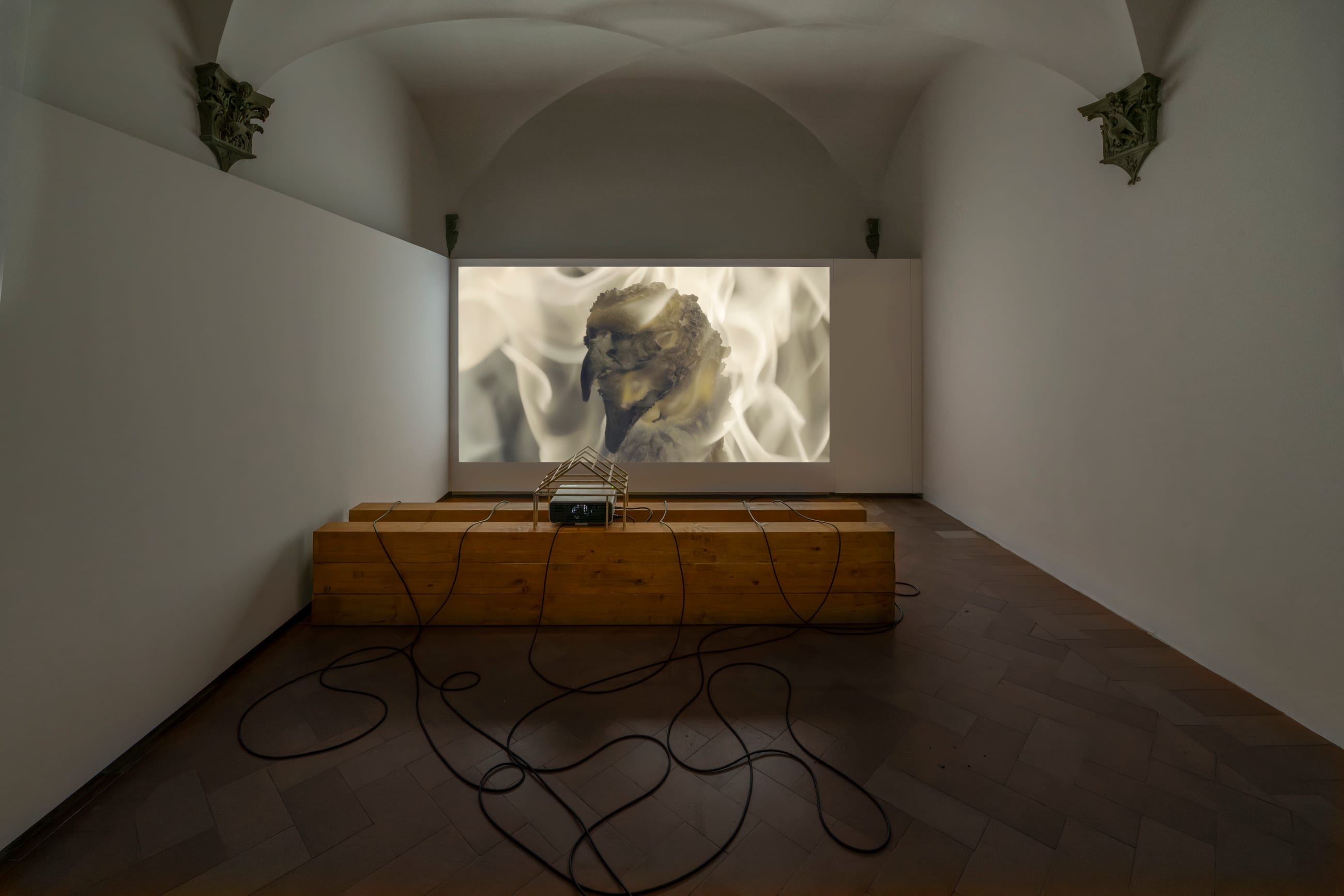 Sala espositiva con una proiezione video e una lunga panca in legno che sostiene il proiettore