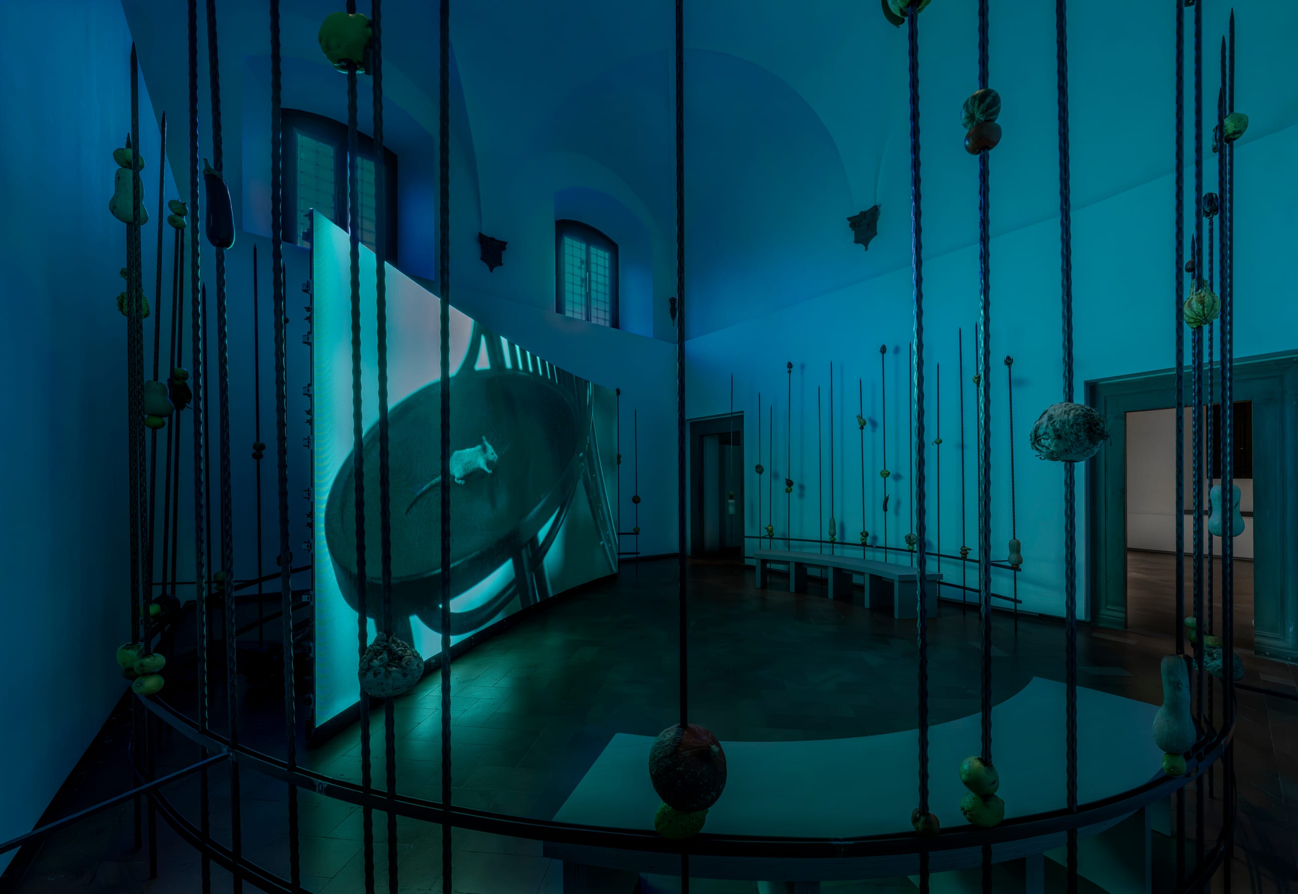 Sala in penombra con aste verticali, oggetti appesi e uno schermo video curvo