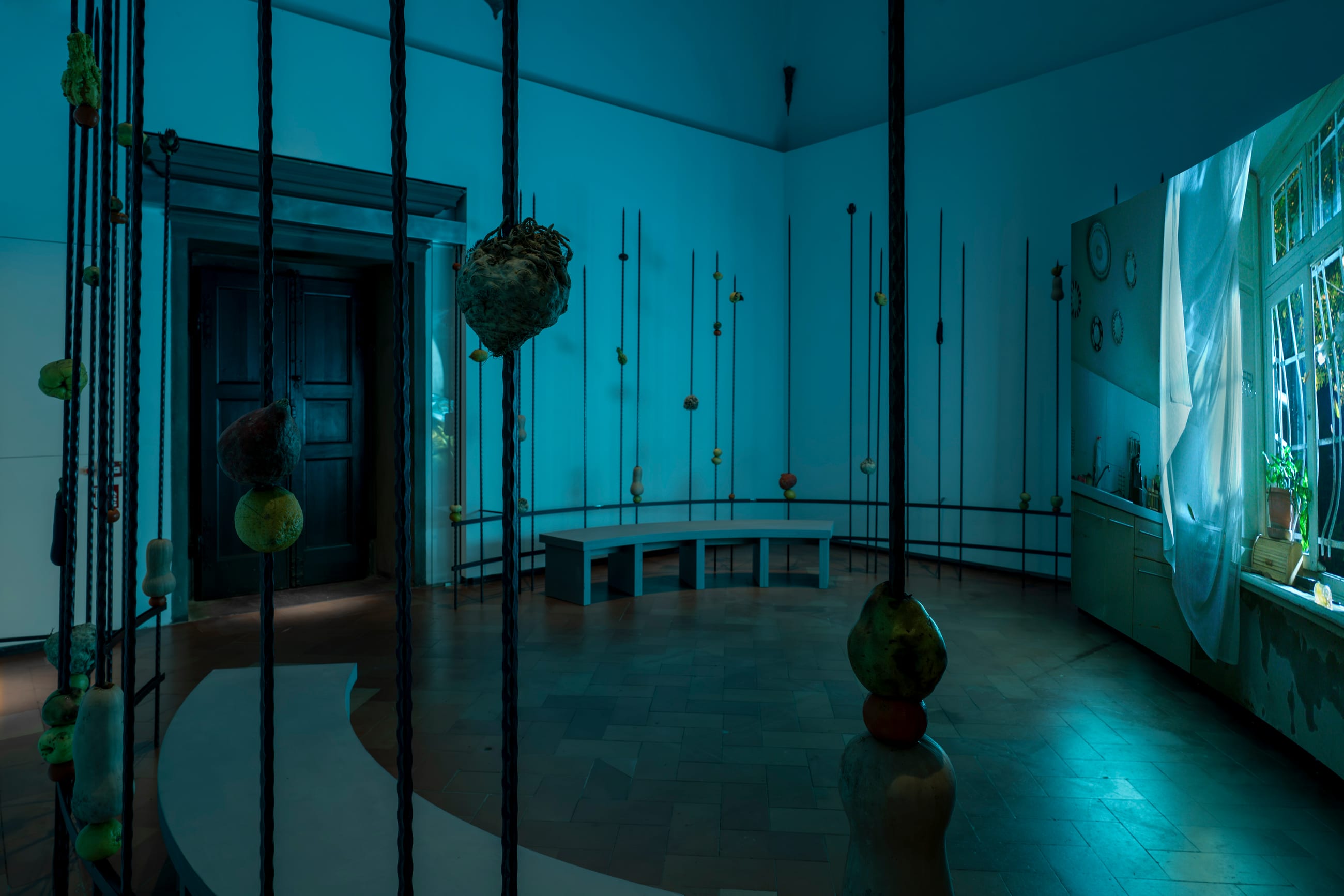 Sala in luce blu con aste verticali, oggetti sospesi e una panca curva