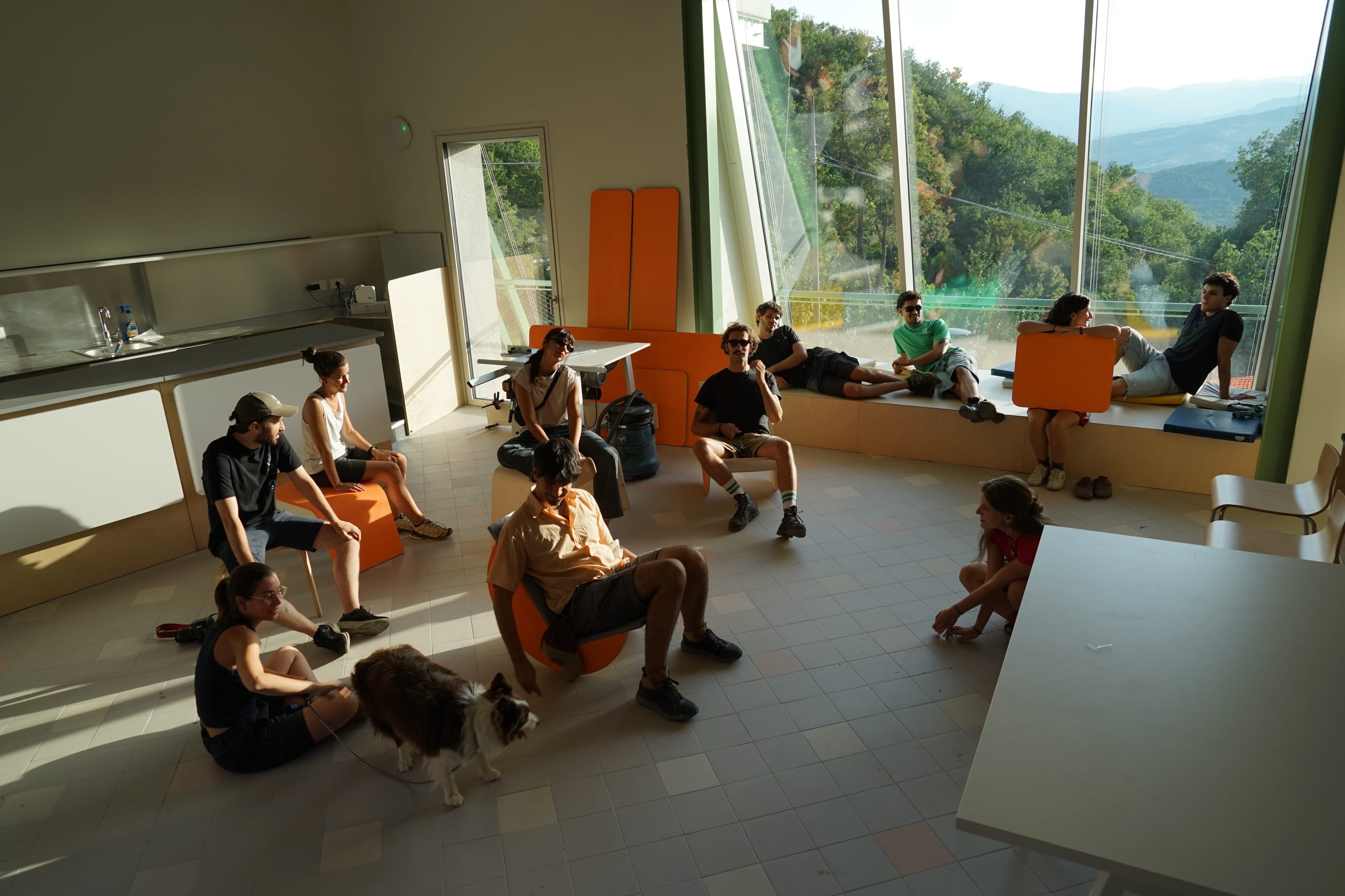 Gruppo seduto su arredi in legno in una sala con grandi vetrate aperte sulle colline
